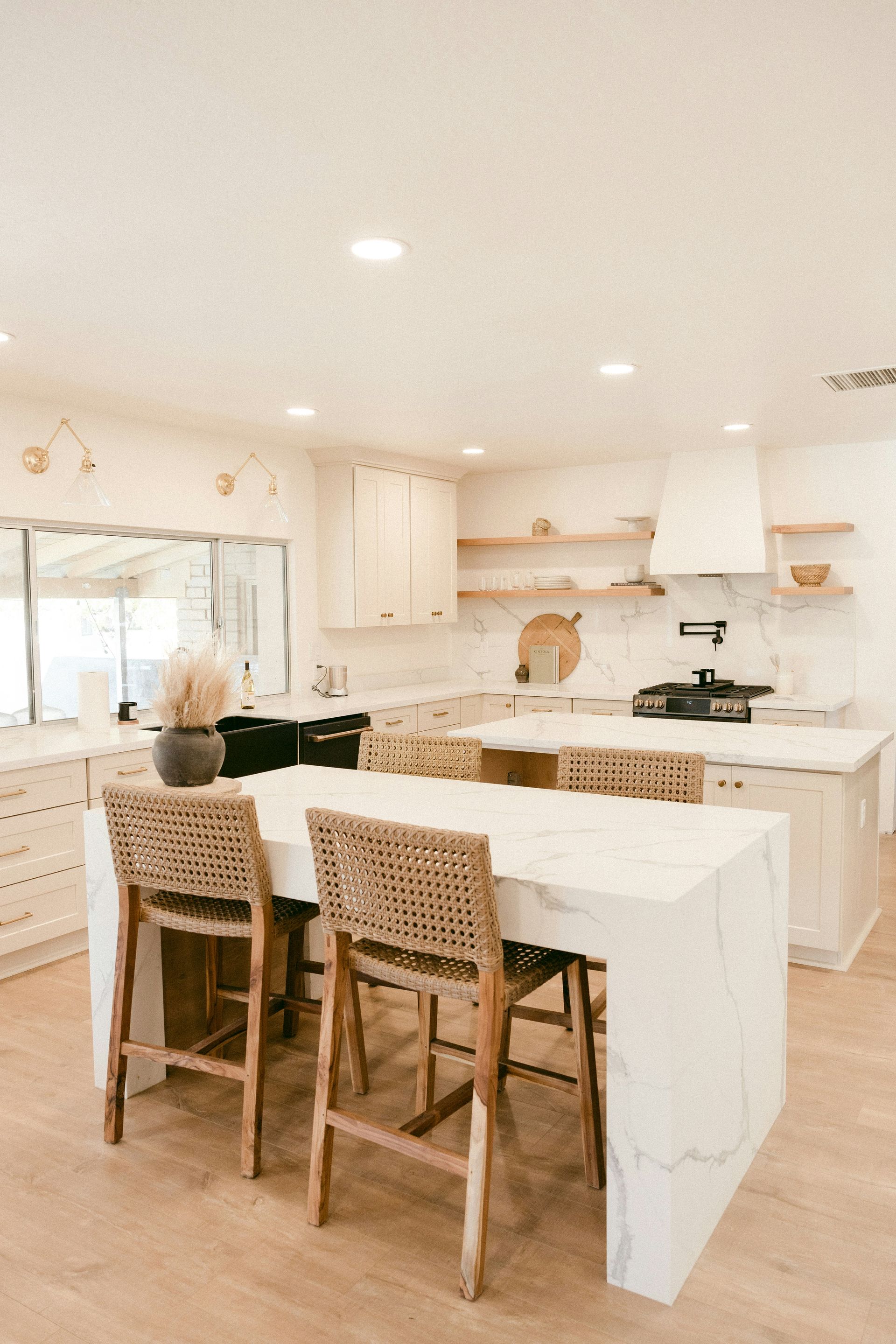 A kitchen with white marble islands, woven bar stools, and light wood flooring